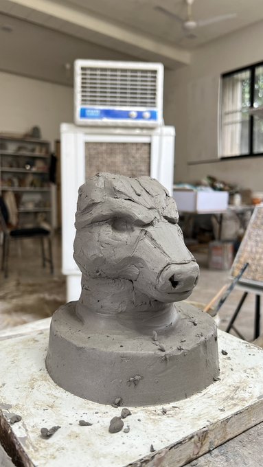 Unfired clay sculpture of a ram or goat head on a wooden base in what appears to be an art studio with an air cooler in the background.