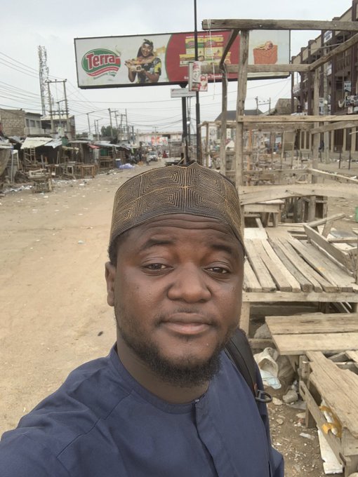 A man wearing a brown patterned kippah and dark blue top takes a selfie outdoors in what appears to be a bustling West African market area, with a "Terra" branded billboard visible in the background. The image captures a personal "GM" (Good Morning) greeting to celebrate his 30-day milestone with RetiumChain, sharing his local surroundings with the community.