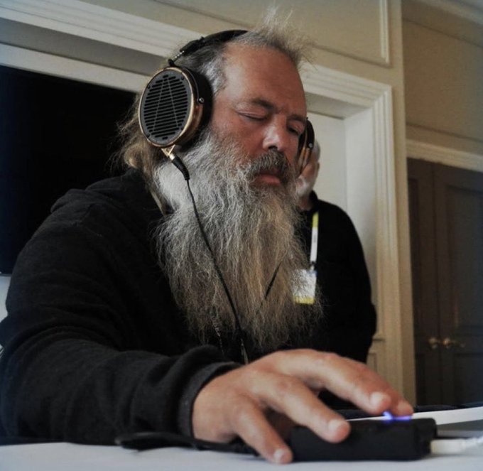 A man with a long gray beard wearing headphones is focused on operating a device with his hand on what appears to be a laptop.