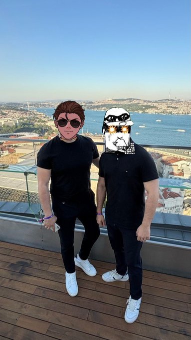 The image shows two men posing for a picture at an outdoor event, possibly a rooftop party or gathering. They are standing next to each other with their faces partially obscured by masks. The background features a beautiful view of the water, adding to the ambiance of the scene. The caption includes various usernames and mentions that the photo was taken on social media platforms like Instagram, Twitter, and Facebook.