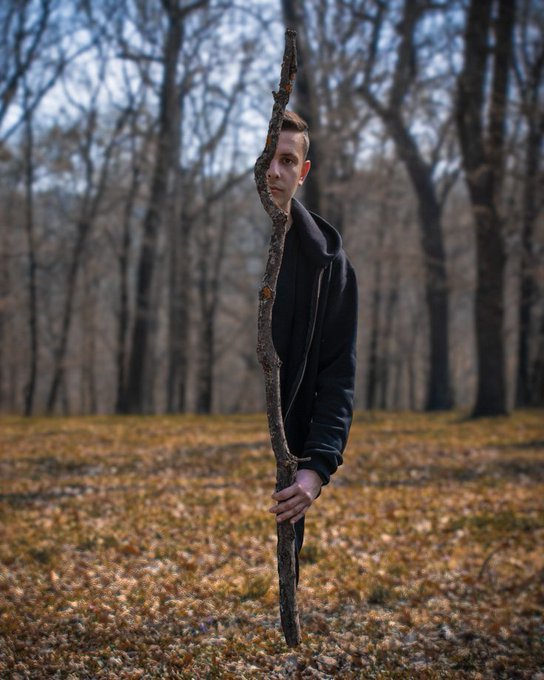 The image features a man standing in a field with his arm wrapped around a tree branch, holding onto it as if he's about to take off into the sky or embark on an adventure. The background of the photo shows trees and a forest setting, creating a sense of being outdoors surrounded by nature. There is also text present in the image that reads "gEvening famo day check-in... how’s the day going so far? stay with @xoobnetwork $RIVER | @RiverdotInc". This suggests that the man might be taking a break from his daily routine to enjoy some time outdoors and appreciate nature.