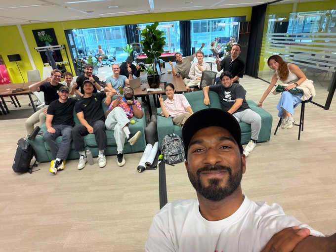 A group of around 15 casually dressed individuals pose together in a modern lounge space featuring green sofas, with one person in the foreground taking a selfie while others smile, gesture, and give thumbs up from the background. The image captures the friendly, collaborative atmosphere of the @pradologue from @goatfishxyz visit, with visible "GOAT" text on one attendee's shirt reflecting the company's branding mentioned in the post about discussing narratives for @MeteoraEco, @JupiterExchange, and @solana.