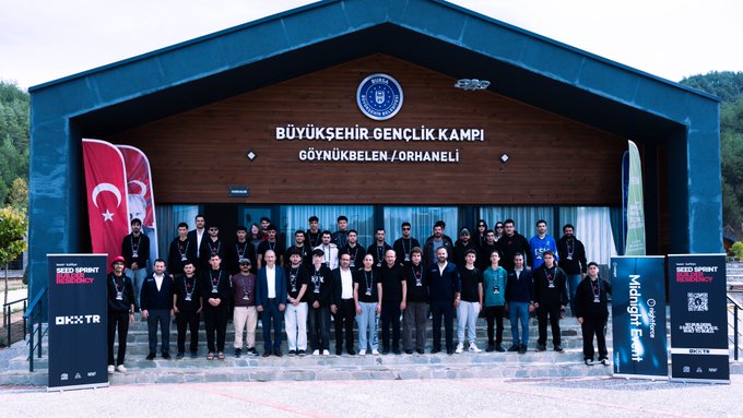 A group of people pose for a photo in front of a wooden building labeled "BÜYÜKŞEHİR GENÇLİK KAMPI GÖYNÜKBELEN / ORHANELİ" with Turkish flags and event banners for "RED SPRINT" and "Midnight Sprint" visible.