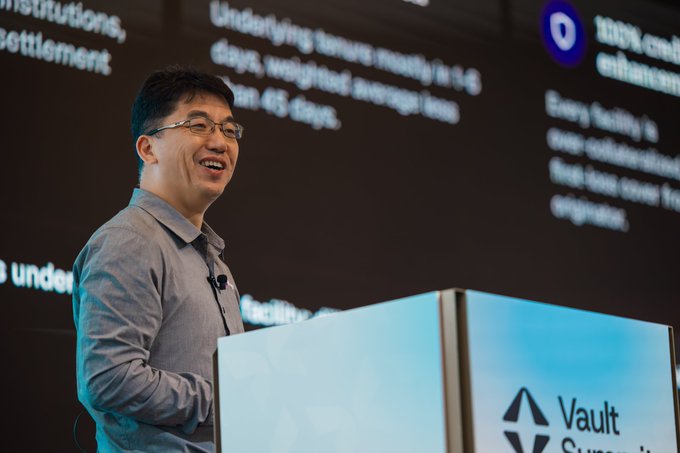 A smiling man with glasses speaks at a podium labeled 'Vault Summit' with a presentation slide about institutions and settlements visible behind him.