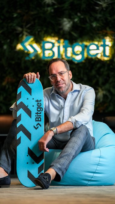 A man wearing glasses and a light blue shirt sits on a beanbag chair holding a teal and black skateboard branded with 'Bitget', with a green plant wall featuring the 'Bitget' logo behind him.