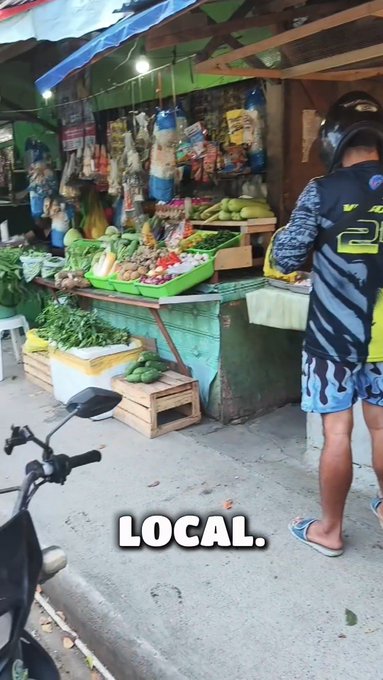 A street market stall selling fresh vegetables and hanging snacks with a man in a helmet and black shirt standing nearby; text 'LOCAL.' overlaid at the bottom.