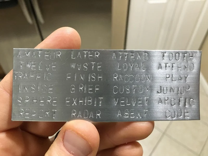 A hand holds a metal seed phrase plate engraved with 24 recovery words (including "AMATEUR," "TWELVE," "TRAFFIC," "INSIDE," "SPHERE," and "REPORT"). The image illustrates the exact security risk the post warns about — publicly sharing a seed phrase plate makes a Bitcoin wallet vulnerable, regardless of whether it depicts a genuine backup, and any phrase exposed this way should be treated as compromised.