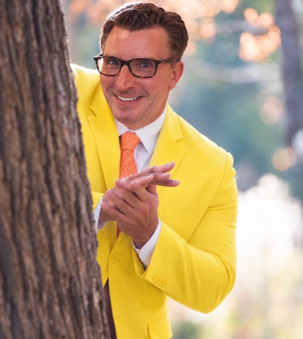 A man in a bright yellow suit and orange tie wearing black glasses smiles while peeking out from behind a tree.