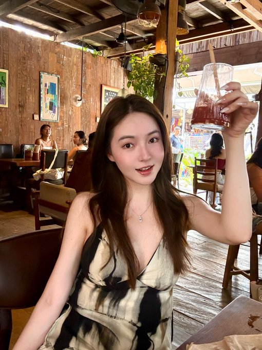 A young woman smiles while raising a glass of dark iced coffee at a rustic wooden café, with other patrons visible in the background—capturing the relaxed, leisurely vibe of "苍古的悠闲时光" (Canggu's leisurely moments) as she invites fellow digital nomads to meet up over coffee.