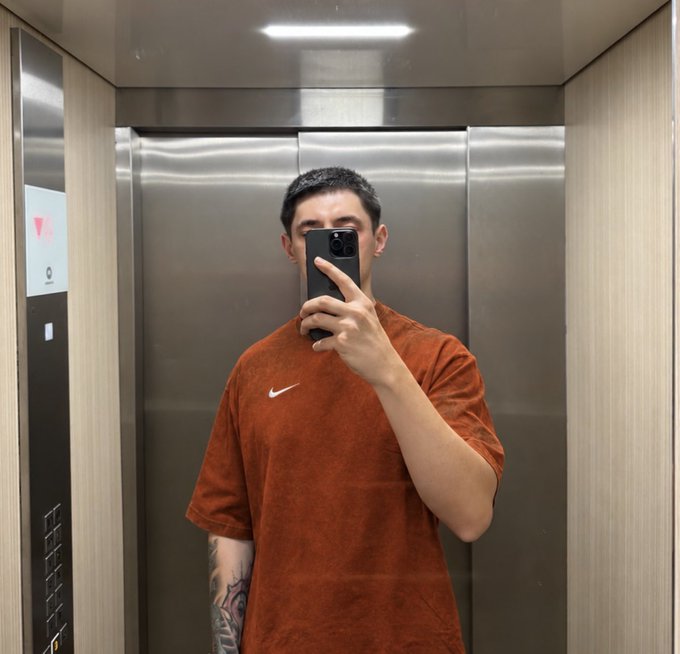 A mirror selfie taken inside an elevator by a young man wearing a rust-orange Nike t-shirt, holding a phone that partially obscures his face.