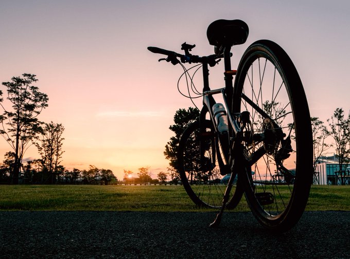 A bicycle is silhouetted against a colorful sunset sky, standing upright on a paved path beside a grassy field.