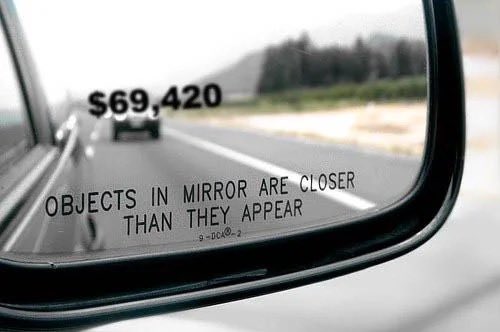 A side-view car mirror showing a vehicle behind displays '$69,420' in bold text overlaid on the mirror's reflection, along with the standard 'OBJECTS IN MIRROR ARE CLOSER THAN THEY APPEAR' warning.