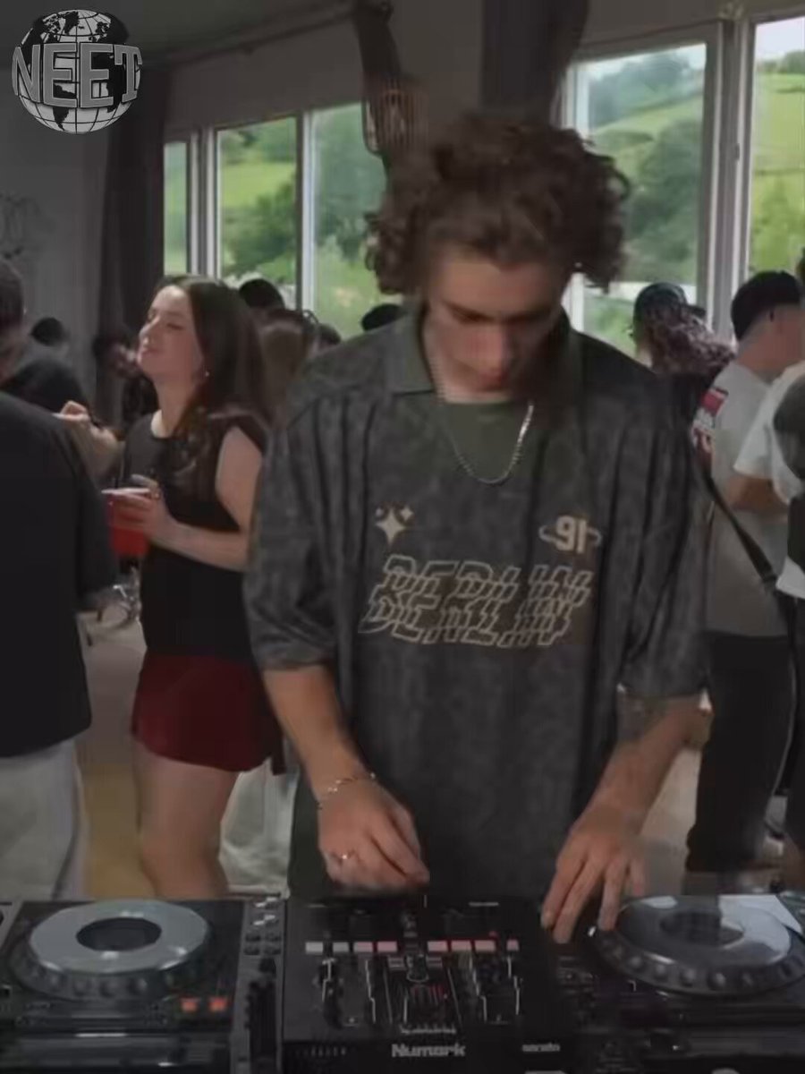 The image features a man standing behind a DJ booth, playing music for an audience. The scene takes place at a party or event where people are gathered around him, enjoying his performance. There are several individuals visible in the background, some of whom appear to be dancing and having fun. A cup can also be seen on one of the tables nearby.