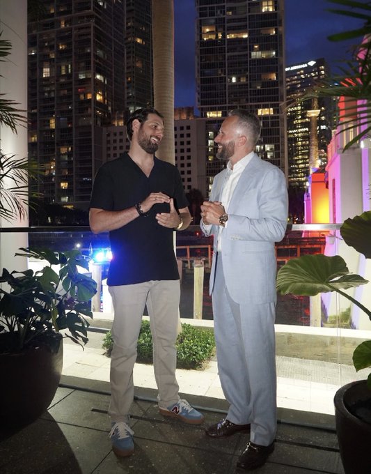 Two men—one in a casual black polo and the other in a light blue suit—share a laugh on a high-rise balcony at night, with a city skyline glowing behind them, matching the post's playful tone about the unique appeal of "Doginal Dogs."