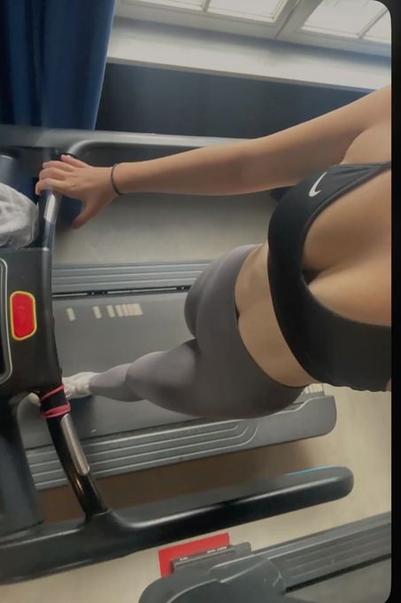 Photo taken inside a gym showing a woman in a black Nike sports bra and gray leggings using an elliptical or stair-climber machine.