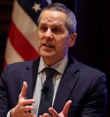 A man in a dark suit and tie speaking while gesturing with his hands, with an American flag visible in the background behind him.