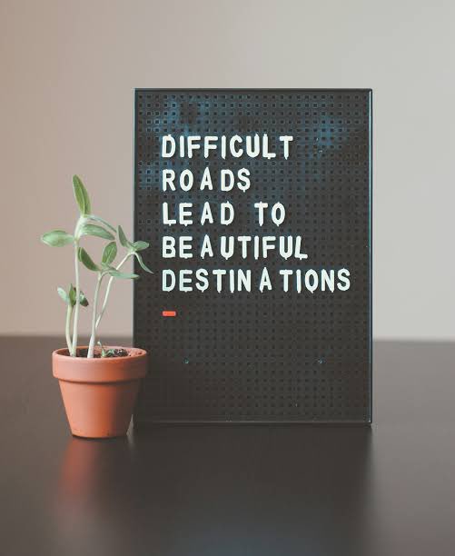 A motivational black pegboard sign reading "DIFFICULT ROADS LEAD TO BEAUTIFUL DESTINATIONS" sits next to a small potted succulent plant on a dark wooden surface.