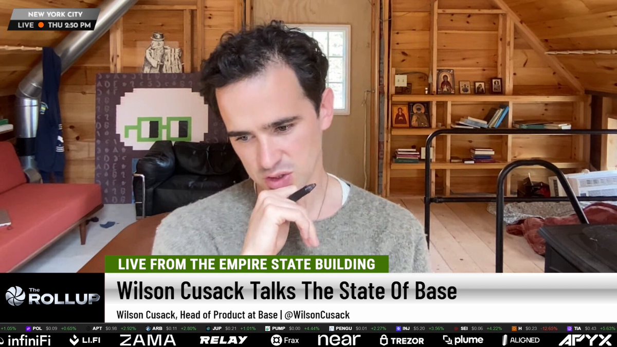 The image shows a man sitting at a desk with his hand on his chin, looking thoughtful or deep in thought. The background of the image features a wooden room with a couch and several bookshelves filled with various items such as books and knick-knacks. There is also an advertisement for Bitcoin displayed prominently in the scene.