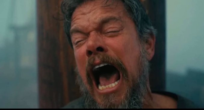 A close-up still from a film showing a man with a beard and messy hair screaming with his eyes closed.