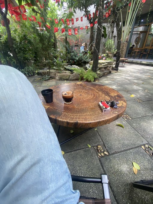 A wooden coffee table with a small cup and ashtray sits on a tiled patio in a courtyard decorated with red flags, viewed past a person in a light blue shirt.
