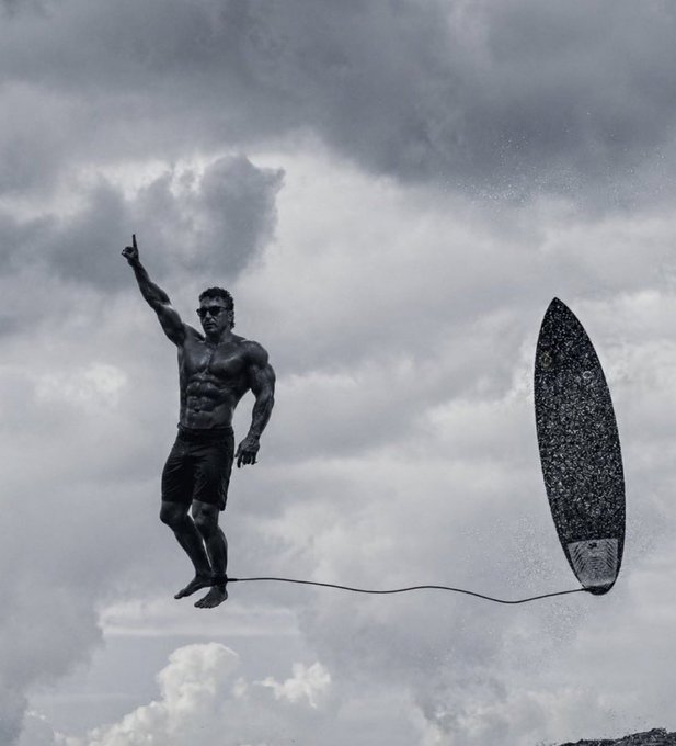 The image features a man standing on a surfboard, with his arms raised high above his head as he looks up at the sky. The background of the photo shows clouds, suggesting that they are taking place during or after a storm. There is also an object in the scene, which appears to be a surfboard, located near the center-right part of the image.