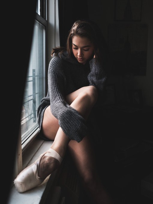 A moody portrait of a young woman in an oversized gray sweater sitting on a window sill wearing pointe shoes, gazing downward.
