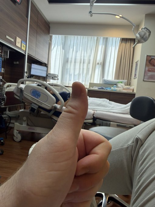 A hand gives a thumbs-up in the foreground of a hospital delivery room, with medical equipment and a hospital bed visible behind. The image humorously parallels the "mainnet first" joke by suggesting a more meaningful "launch" — the birth of a baby — alongside the sterile, technical setting of a hospital.