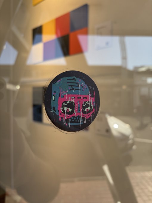 A circular sticker featuring a teal and pink graffiti-style skull design attached to a glass surface.