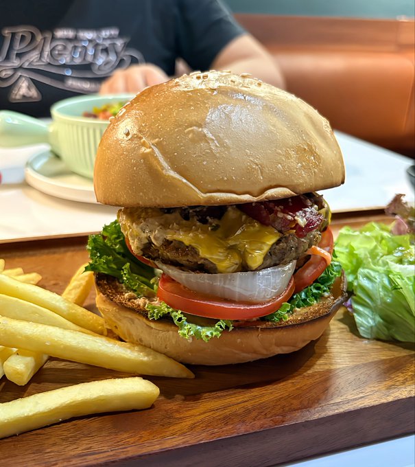 A large cheeseburger with lettuce, tomato, onion, and bacon served with french fries on a wooden board at what appears to be a Plenty restaurant.