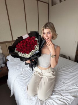 A smiling woman with short blonde hair sits on a bed holding a large bouquet of red roses wrapped in black paper.