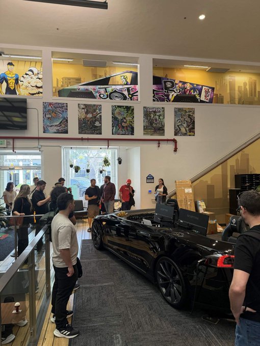 Interior of what appears to be a comic book or collectibles store/gallery with people gathered around a black sports car parked inside.