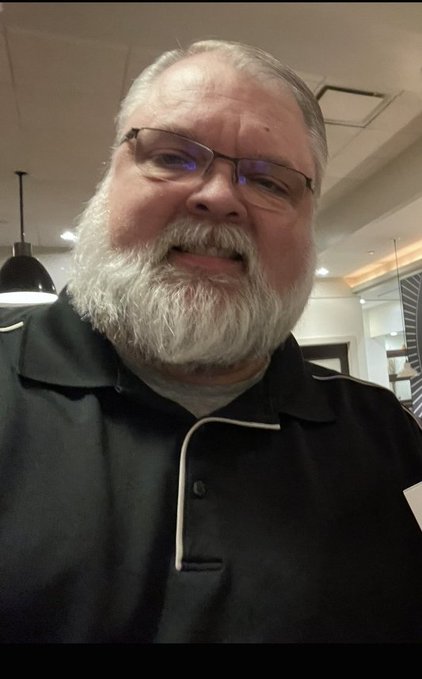 A close-up selfie of an older man with a white beard and glasses wearing a black polo shirt, taken indoors.