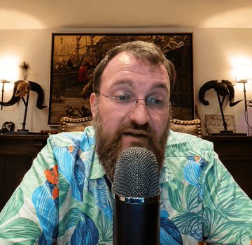 A man with a goatee and a large goatee wearing glasses , sits behind a microphone talking into a podcast mic while holding his hand up to the camera for the photo. He's dressed casually, wearing a shirt with palm trees on it, suggesting he is in an outdoor setting or possibly a tropical location. The image also features a photo of a painting and two lamps mounted at different heights above him. A caption below the photo reads "BEARISH: Cardano'S native token has fallen below the $0.20 level for the first time in more than five years, adding to a difficult period for the blockchain ecosystem". The main focus of the image is on the man and his position behind the microphone.