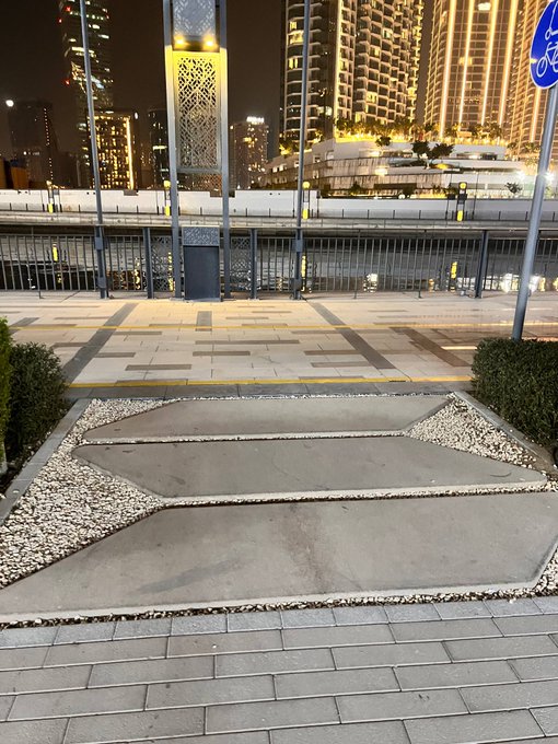 A nighttime urban plaza with stone pathways, decorative gravel landscaping, and illuminated high-rise buildings in the background.