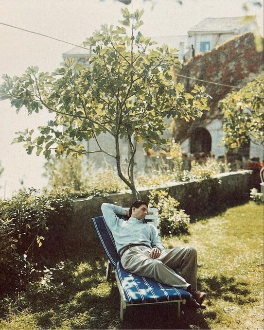 A vintage-style photo of a man relaxing on a blue striped lounge chair in a garden, leaning back under a tree.