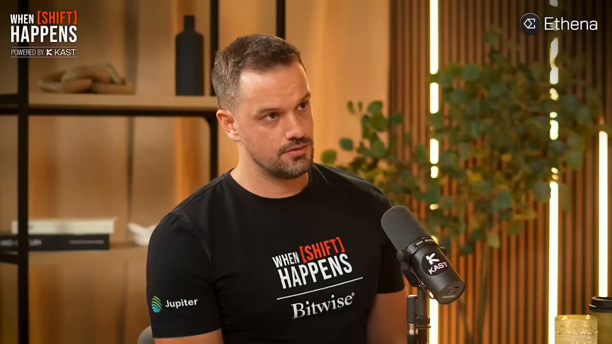 The image features a man wearing a black t-shirt with white text that says "Hands off! Here comes the bear market" while sitting at a desk or table, possibly in an office setting. The background of the image shows a bookshelf filled with various items such as bottles and books.