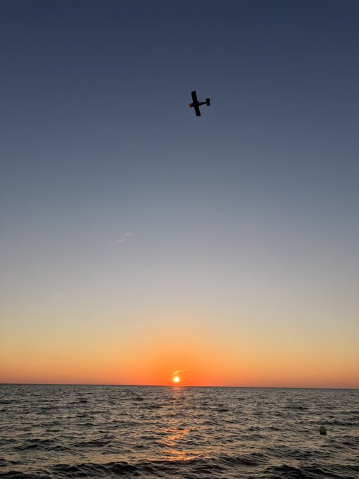 A silhouetted small aircraft flying over the ocean against a vibrant sunset sky with the sun dipping toward the horizon.