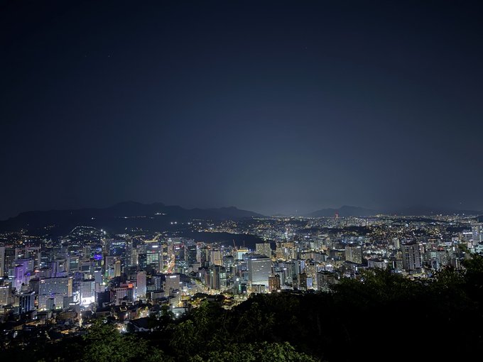 A nighttime cityscape of a brightly lit urban skyline with a river winding through the city and dark mountains in the background.