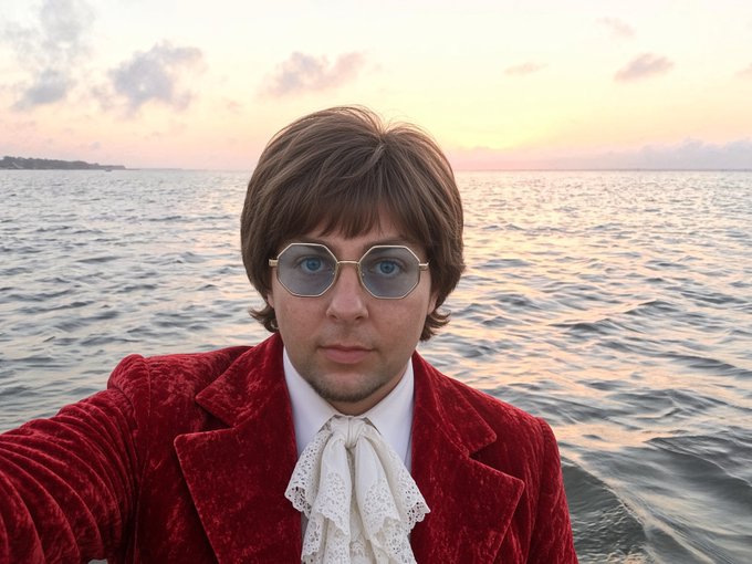 A man dressed in a red velvet jacket and white ruffled shirt takes a selfie, wearing tinted round glasses and a brown wig resembling John Lennon, with the sea and a sunset sky behind him.