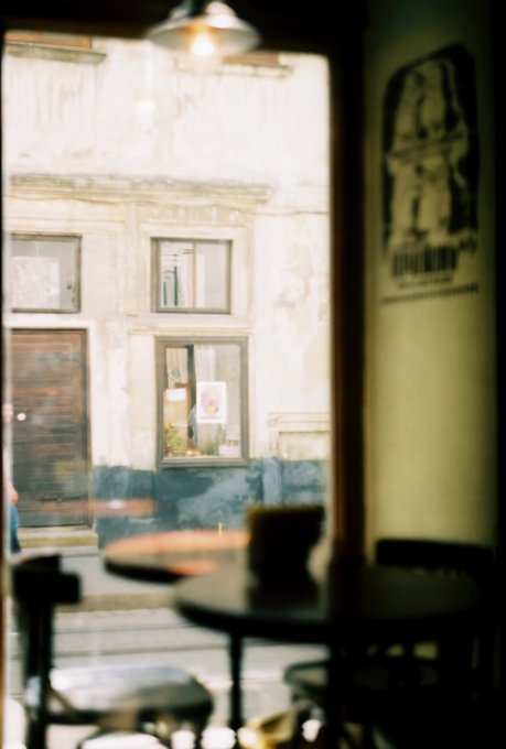 A view through a window shows an old, weathered building across the street. In the foreground, out-of-focus tables and chairs suggest the photo was taken from inside a cafe.
