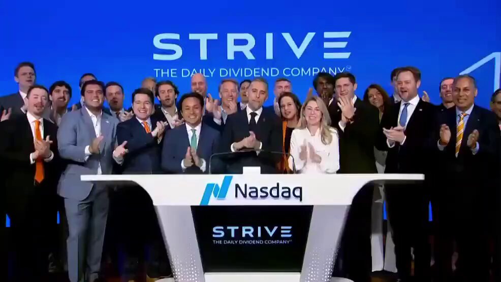 The image shows a group of people standing around a table, clapping their hands together. The people are dressed in business attire, with some wearing ties. They appear to be celebrating an event or achievement related to Stripe, a company that provides payment processing services. The group is gathered behind a desk, and there's a large screen displaying the Stripe logo on it. The image also includes text about the company's daily dividends and their estimated value in Bitcoin.