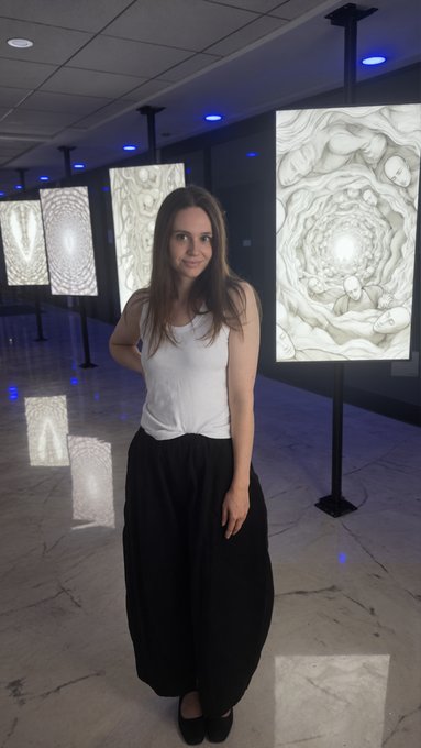 A woman with long brown hair stands in an art gallery, wearing a white sleeveless top and black wide-leg pants, with illuminated textured artworks displayed behind her.