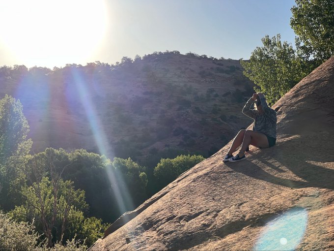 A person sits on a large sandstone rock formation, with sunlight streaming through a canyon landscape dotted with trees.
