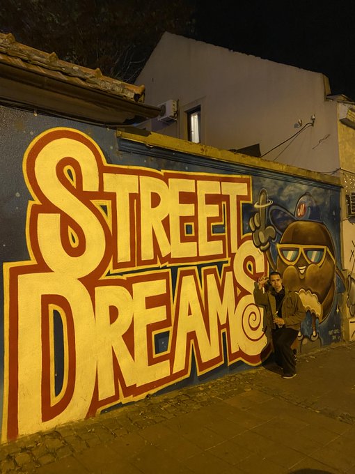 A person poses in front of a vibrant "Street Dreams" mural featuring an egg-shaped character wearing a cap and sunglasses, with the "ST" initials cleverly worked into the artwork—matching the post about volunteering at the ST Balkan Booth for Solana Summit Belgrade.