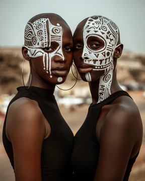 Two women in black tank tops stand back-to-back, their faces adorned with elaborate black and white tribal paint designs, wearing large hoop earrings.