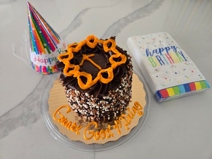 A chocolate birthday cake decorated with orange frosting reading 'Good Morning' and 'Happy Birthday' sits next to a party hat and a 'happy BIRTHDAY' gift card on a marble countertop.