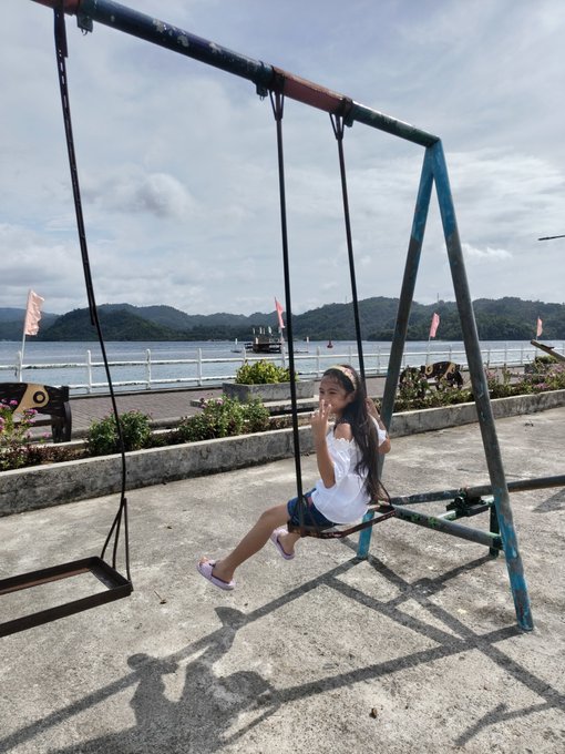 Photograph of a smiling young girl in a white top and denim shorts swinging on an old metal swing set at a seaside promenade, with water, boats, green hills, and a partly cloudy sky in the background.