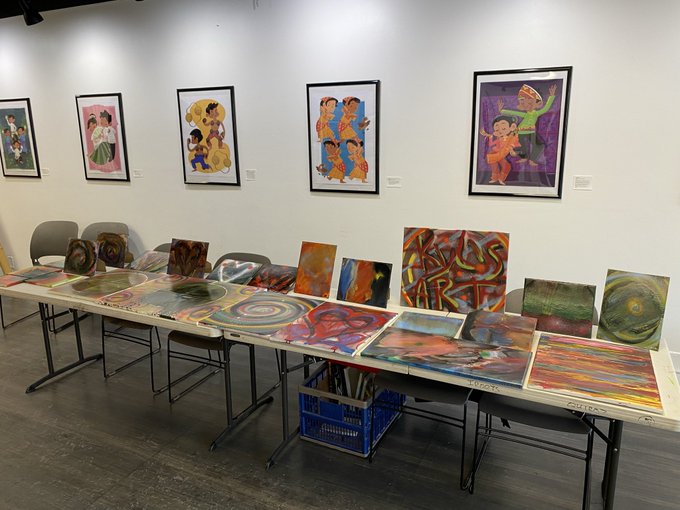 An art gallery display shows colorful framed paintings hung on a white wall above a long table displaying numerous smaller, abstract and figurative artworks.