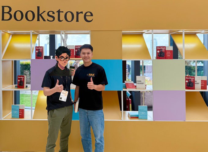 A photo of two people posing with thumbs up in front of a bookstore display, with one person's face covered by an anime-style avatar; the books on the shelves are titled "FiberMark" with a "Bookstore" header visible.