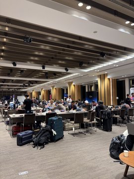 A bustling conference hall filled with hackers and developers working at long tables, surrounded by laptops and backpacks, as a large screen displays "ETHGlobal" in the background. The image reflects the intense atmosphere of day 2 at the ETHGlobal Tokyo hackathon.